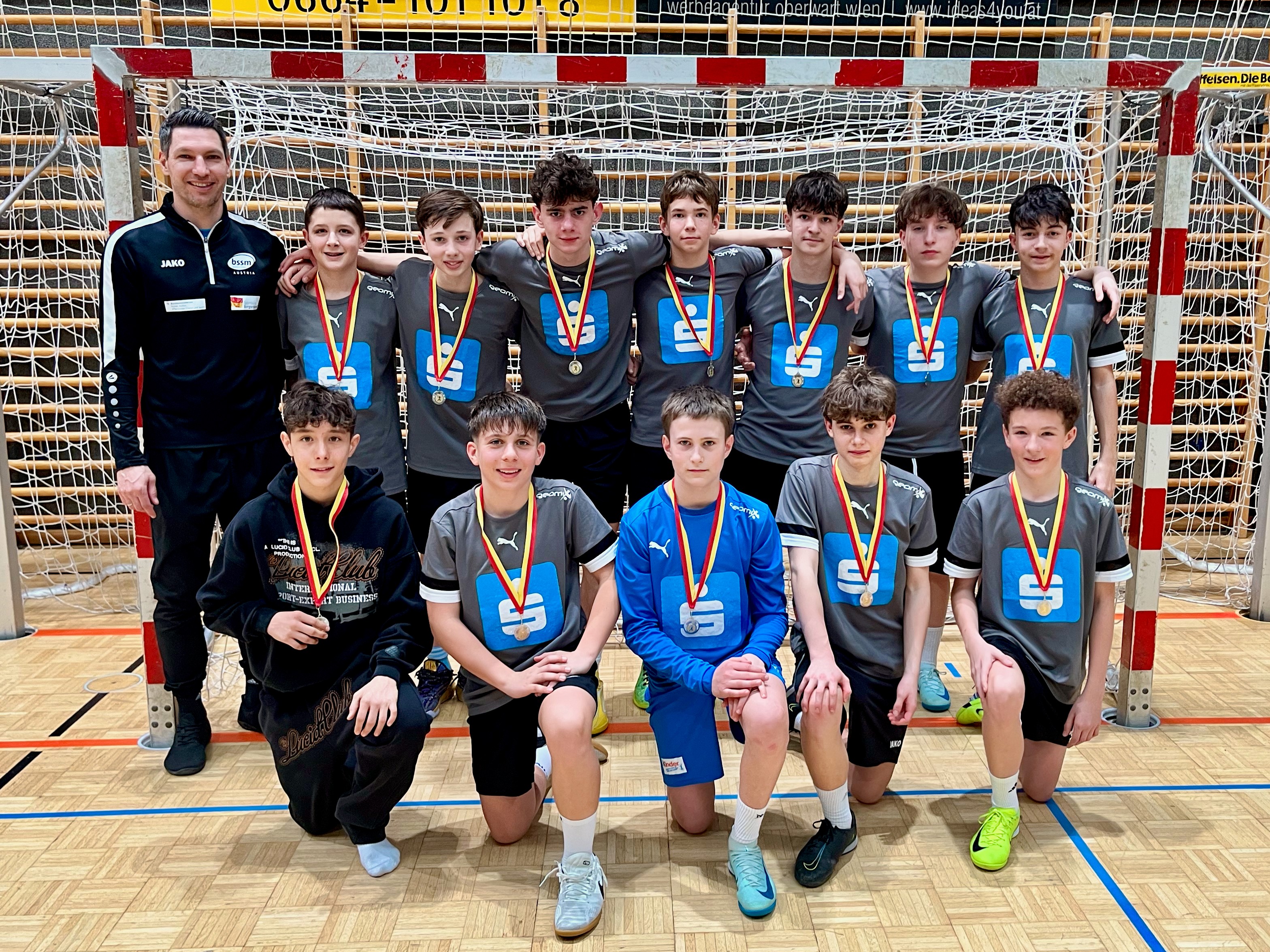 Das Foto zeigt eine Jugend-Handballmannschaft in einer Sporthalle. Die Gruppe steht vor einem Handballtor mit Netz. Insgesamt sind elf Spieler und ein Trainer zu sehen.  Der Trainer steht links neben der Mannschaft und trägt einen dunklen Trainingsanzug. Hinter ihm stehen sieben Spieler Schulter an Schulter vor dem Tor. Vor ihnen knien vier weitere Spieler. Alle Jugendlichen tragen graue Trikots mit einem großen blauen Logo auf der Brust sowie schwarze Sporthosen. Ein Spieler in der vorderen Reihe trägt ein blaues Torwarttrikot.  Alle Spieler haben Medaillen mit rot-gelben Bändern um den Hals, was darauf hindeutet, dass sie bei einem Turnier erfolgreich waren. Die meisten lächeln in die Kamera. Der Hallenboden aus Holz und die Linien des Spielfelds sind deutlich zu sehen.  Das Foto vermittelt einen stolzen und freudigen Moment nach einem sportlichen Erfolg der Mannschaft.