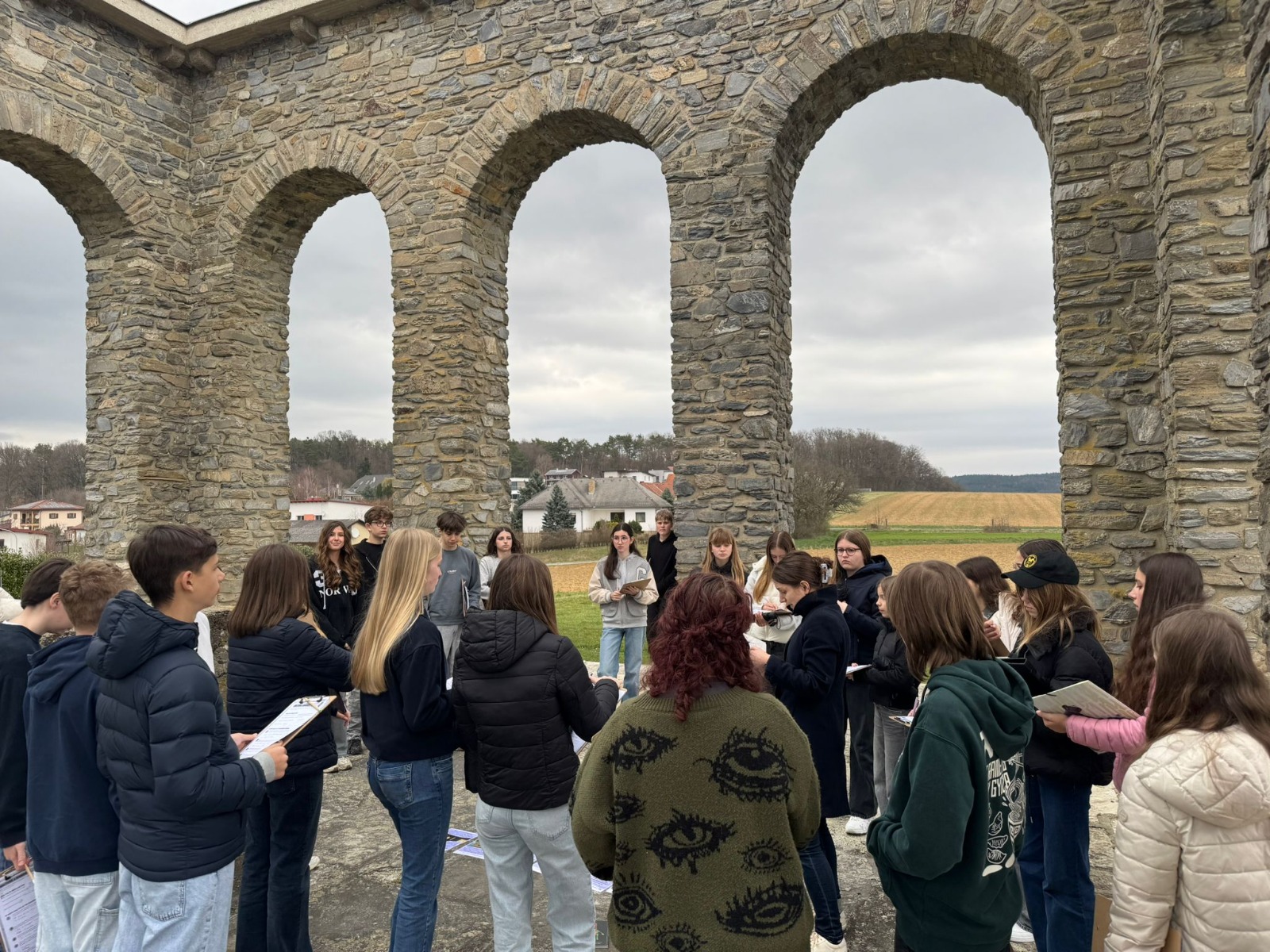 Das Bild zeigt eine Gruppe von Jugendlichen, die sich im Freien unter einer alten Steinstruktur mit mehreren großen Rundbögen versammelt haben. Die Bögen bestehen aus grob behauenen Steinen und erinnern an ein historisches Bauwerk oder ein Denkmal.  Die Jugendlichen stehen in einem Kreis zusammen und halten teilweise Notizblöcke oder Blätter in der Hand, was darauf hindeutet, dass sie an einer gemeinsamen Aktivität teilnehmen – möglicherweise einem Unterricht im Freien, einer Führung oder einer Projektarbeit. Eine Person in der Mitte scheint etwas zu erklären oder vorzulesen, während die anderen aufmerksam zuhören.  Im Hintergrund sind ein paar Häuser, Bäume und Felder zu sehen, was auf eine ländliche Umgebung schließen lässt. Der Himmel ist bewölkt, wodurch eine eher ruhige, etwas kühle Stimmung entsteht.  Insgesamt vermittelt das Bild den Eindruck einer schulischen oder pädagogischen Situation an einem historischen Ort in der Natur.