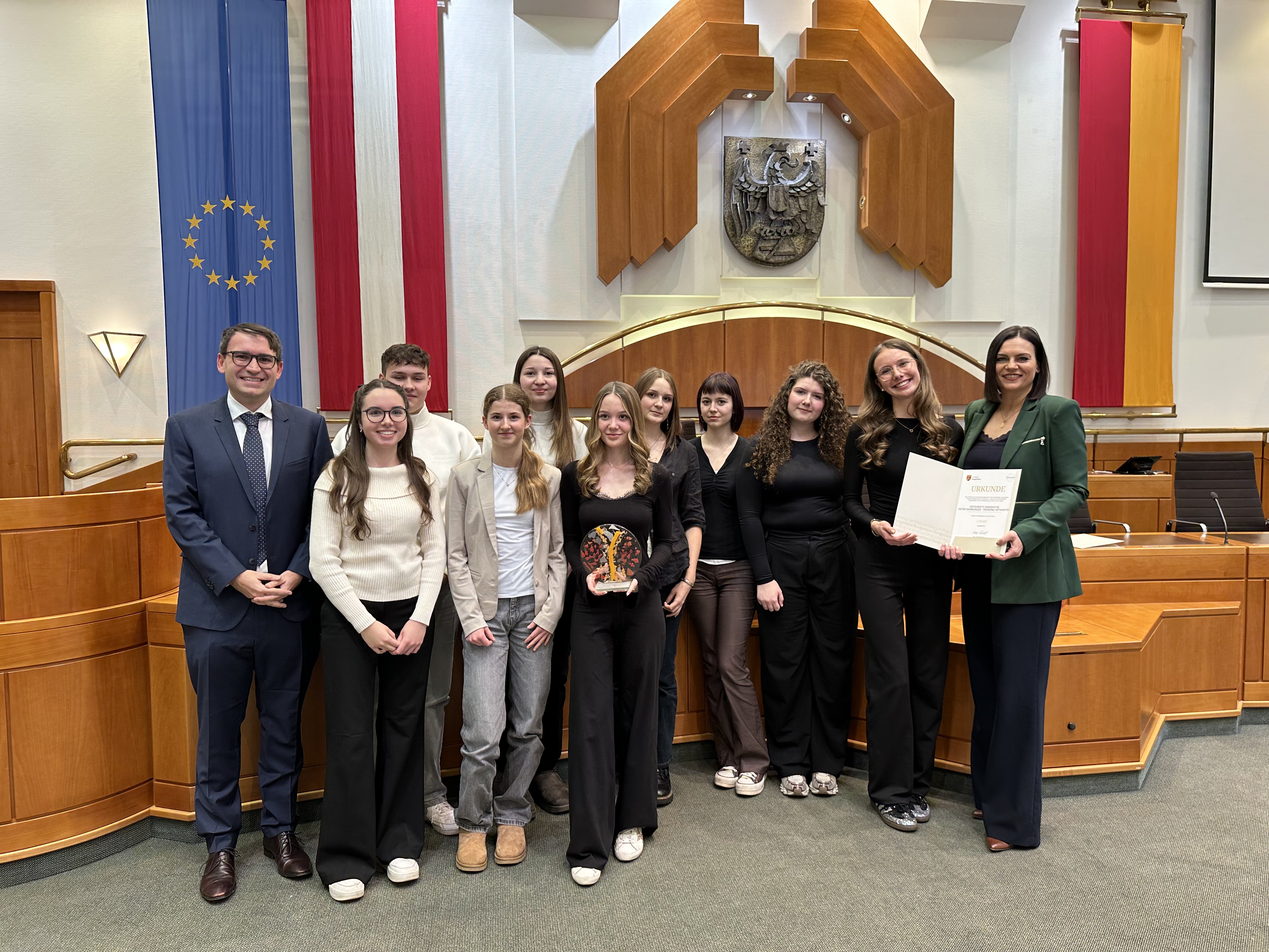 Das Bild zeigt eine Gruppe von Jugendlichen und zwei erwachsenen Frauen sowie einen Mann in einem offiziellen, repräsentativen Raum, der wie ein Sitzungssaal oder Parlamentssaal wirkt. Im Hintergrund hängen große Fahnen, darunter eine blaue Flagge mit gelben Sternen (die Europaflagge) sowie rot-weiß-rote Banner. Über der Gruppe ist ein Wappen angebracht, und der Raum ist mit viel Holz und klaren, symmetrischen Formen gestaltet.  Die Gruppe steht nebeneinander und schaut in die Kamera. Die Jugendlichen sind überwiegend in dunkler oder schlichter Kleidung gekleidet, einige tragen schwarze Oberteile, andere Kombinationen aus hellen und dunklen Kleidungsstücken. In der Mitte hält eine junge Frau eine dekorative Auszeichnung oder Trophäe. Rechts daneben halten zwei erwachsene Frauen gemeinsam eine Urkunde, die auf eine Preisverleihung oder Ehrung hindeutet.  Der Mann auf der linken Seite trägt einen Anzug mit Krawatte und lächelt freundlich. Die beiden Frauen rechts sind ebenfalls elegant gekleidet, eine in einem dunklen Outfit, die andere mit einem grünen Blazer. Insgesamt vermittelt das Bild den Eindruck eines feierlichen Anlasses, vermutlich einer Preisverleihung oder offiziellen Ehrung einer Schülergruppe.