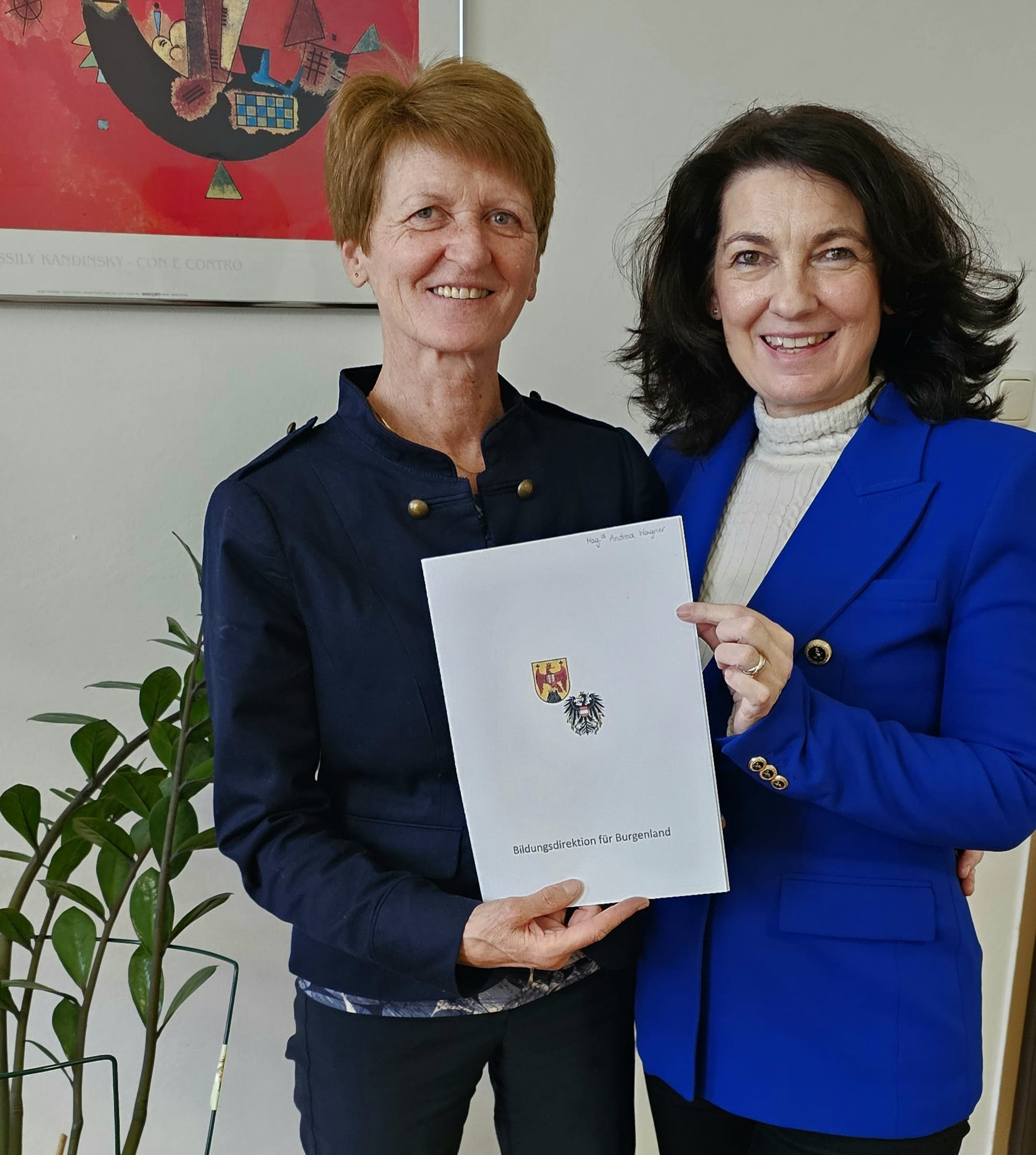 Das Bild zeigt zwei Frauen, die nebeneinander stehen und freundlich in die Kamera lächeln. Sie befinden sich in einem Innenraum, vermutlich in einem Büro oder offiziellen Umfeld.  Die Frau links hat kurzes, hellbraunes Haar und trägt eine dunkelblaue Jacke mit dekorativen Knöpfen. Die Frau rechts hat schulterlanges, dunkles Haar und ist in einem auffälligen blauen Blazer sowie einem hellen Rollkragenpullover gekleidet.  Gemeinsam halten sie eine weiße Mappe oder Urkunde, auf der ein Wappen sowie ein Adler abgebildet sind. Unten ist der Schriftzug „Bildungsdirektion für Burgenland“ zu erkennen, was darauf hindeutet, dass es sich um eine offizielle Auszeichnung oder ein Dokument aus dem Bildungsbereich handelt.  Im Hintergrund hängt ein farbenfrohes, abstraktes Kunstwerk an der Wand, und links ist eine grüne Zimmerpflanze zu sehen. Insgesamt vermittelt das Bild den Eindruck eines feierlichen oder formellen Anlasses, möglicherweise einer Übergabe oder Ehrung.