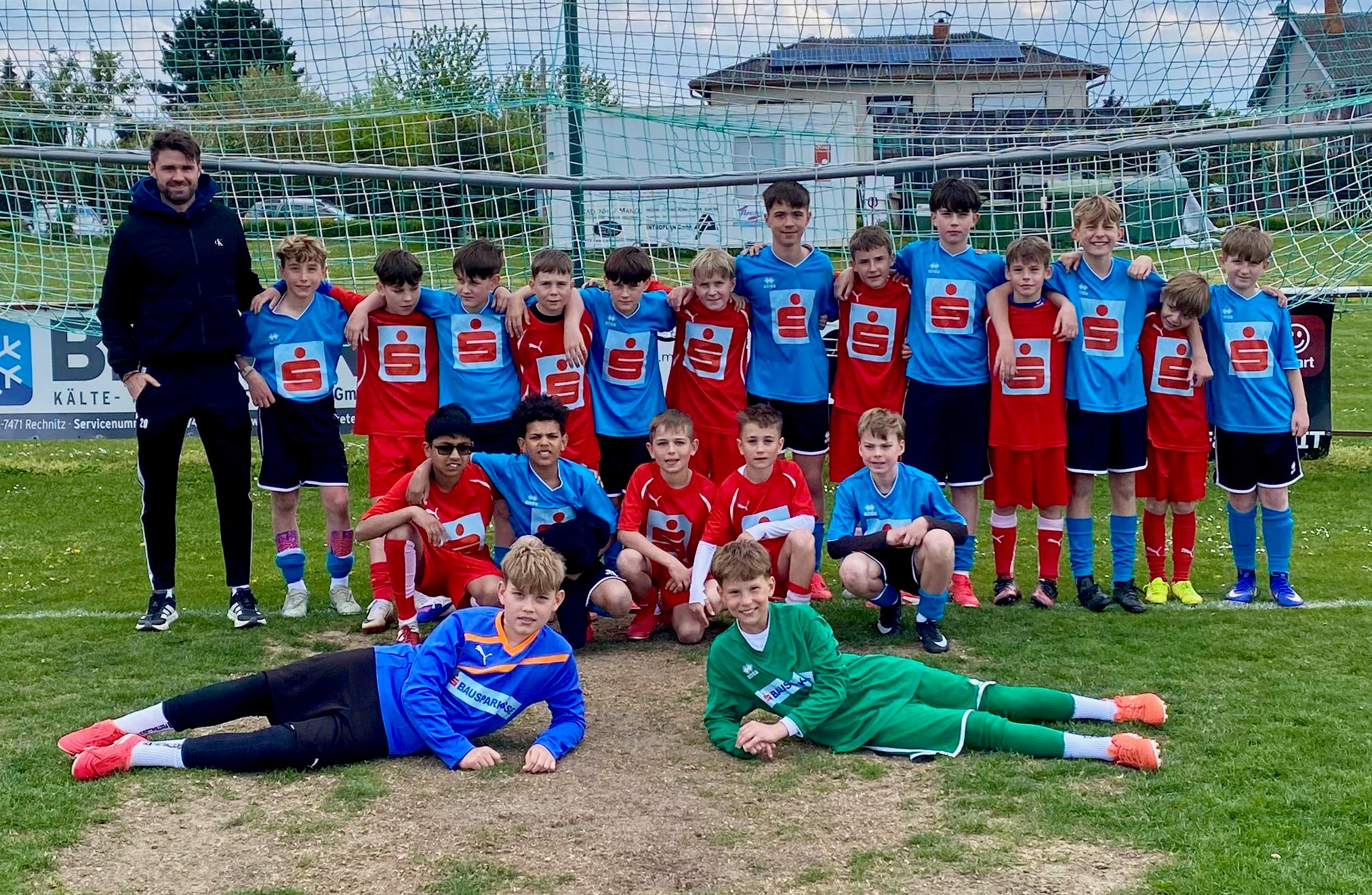 Das Bild zeigt eine Jugendfußballmannschaft, die gemeinsam auf einem Fußballplatz für ein Gruppenfoto posiert. Im Hintergrund ist ein Tor mit Netz zu sehen, dahinter stehen einige Häuser und Bäume.  Die Kinder tragen zwei unterschiedliche Trikotsätze: Ein Teil der Mannschaft ist in roten Trikots gekleidet, der andere in hellblauen. Die Spieler stehen größtenteils in einer Reihe und haben die Arme freundschaftlich umeinander gelegt. Vor ihnen knien einige Kinder, während zwei Jungen in Torwartkleidung ganz vorne auf dem Boden liegen und sich auf die Seite stützen.  Links am Rand steht ein erwachsener Mann, vermutlich der Trainer, der ebenfalls lächelt. Die Stimmung wirkt insgesamt fröhlich und kameradschaftlich, als hätte die Mannschaft gerade ein Spiel hinter sich oder bereite sich darauf vor.  Der Rasen ist leicht abgenutzt, besonders im Vordergrund, was darauf hindeutet, dass der Platz häufig genutzt wird. Insgesamt vermittelt das Bild Teamgeist, Zusammenhalt und Spaß am Sport.