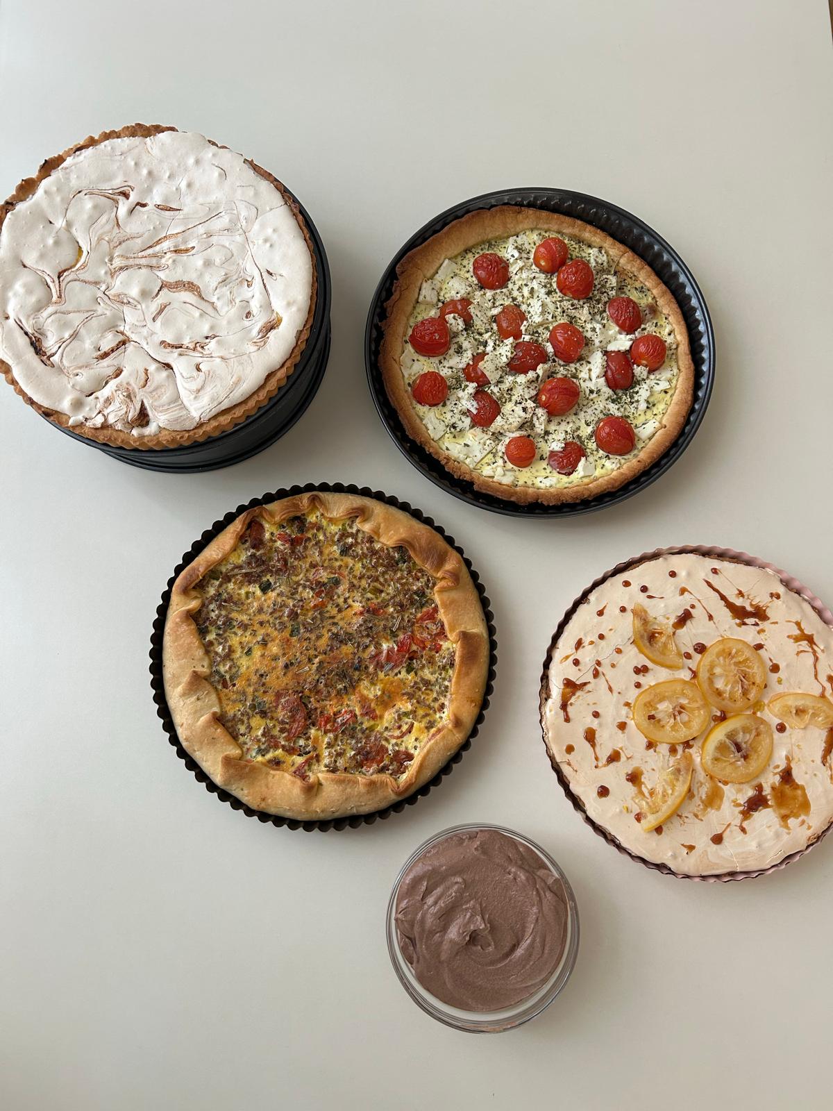 Das Bild zeigt eine Draufsicht auf mehrere Speisen, die auf einer hellen, schlichten Oberfläche angerichtet sind. Insgesamt sind vier runde Kuchen bzw. herzhafte Tartes sowie eine kleine Schale zu sehen, die symmetrisch angeordnet sind.  Oben links befindet sich ein Kuchen mit einer hellen, cremigen Oberfläche, die mit marmorierten braunen Mustern verziert ist. Daneben, oben rechts, liegt eine herzhafte Tarte mit goldbraunem Rand, belegt mit kleinen roten Tomaten und weißen Käsewürfeln, die mit Kräutern bestreut sind.  Unten links sieht man eine weitere herzhafte Tarte mit gefaltetem Rand. Sie ist reichlich gefüllt, vermutlich mit einer Mischung aus Fleisch oder Gemüse und Ei, und ebenfalls leicht gebräunt. Unten rechts befindet sich ein Kuchen mit heller Creme, dekoriert mit dünnen Zitronenscheiben und Karamell- oder Sirupstreifen.  In der Mitte unten steht eine kleine Schüssel mit einer glatten, schokoladenbraunen Creme. Insgesamt wirkt die Anordnung ordentlich und appetitlich, mit einer Mischung aus süßen und herzhaften Speisen.