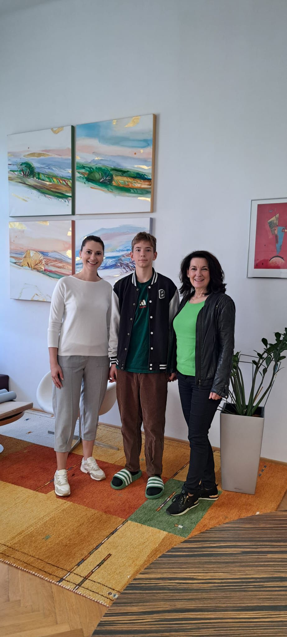 Das Bild zeigt drei Personen, die nebeneinander in einem hellen, modern eingerichteten Raum stehen und in die Kamera lächeln. In der Mitte steht ein Jugendlicher mit kurzen Haaren, der eine dunkle Jacke über einem grünen Shirt sowie braune Hose und grüne Hausschuhe trägt. Links von ihm steht eine Frau mit zusammengebundenen Haaren, die ein weißes Oberteil, eine helle Hose und weiße Sneakers trägt. Rechts steht eine weitere Frau mit dunklen, schulterlangen Haaren, die ein grünes Shirt, eine schwarze Jacke und dunkle Hose mit schwarzen Schuhen trägt.  Der Raum wirkt wohnlich und stilvoll: An der weißen Wand hängen mehrere farbenfrohe, abstrakte Landschaftsbilder in sanften Pastell- und Grüntönen. Rechts steht eine große Zimmerpflanze in einem weißen Topf, und links ist ein moderner Stuhl zu sehen. Auf dem Boden liegt ein großer Teppich mit geometrischem Muster in warmen Farben wie Orange, Gelb und Grün. Insgesamt vermittelt das Bild eine freundliche, entspannte Atmosphäre.