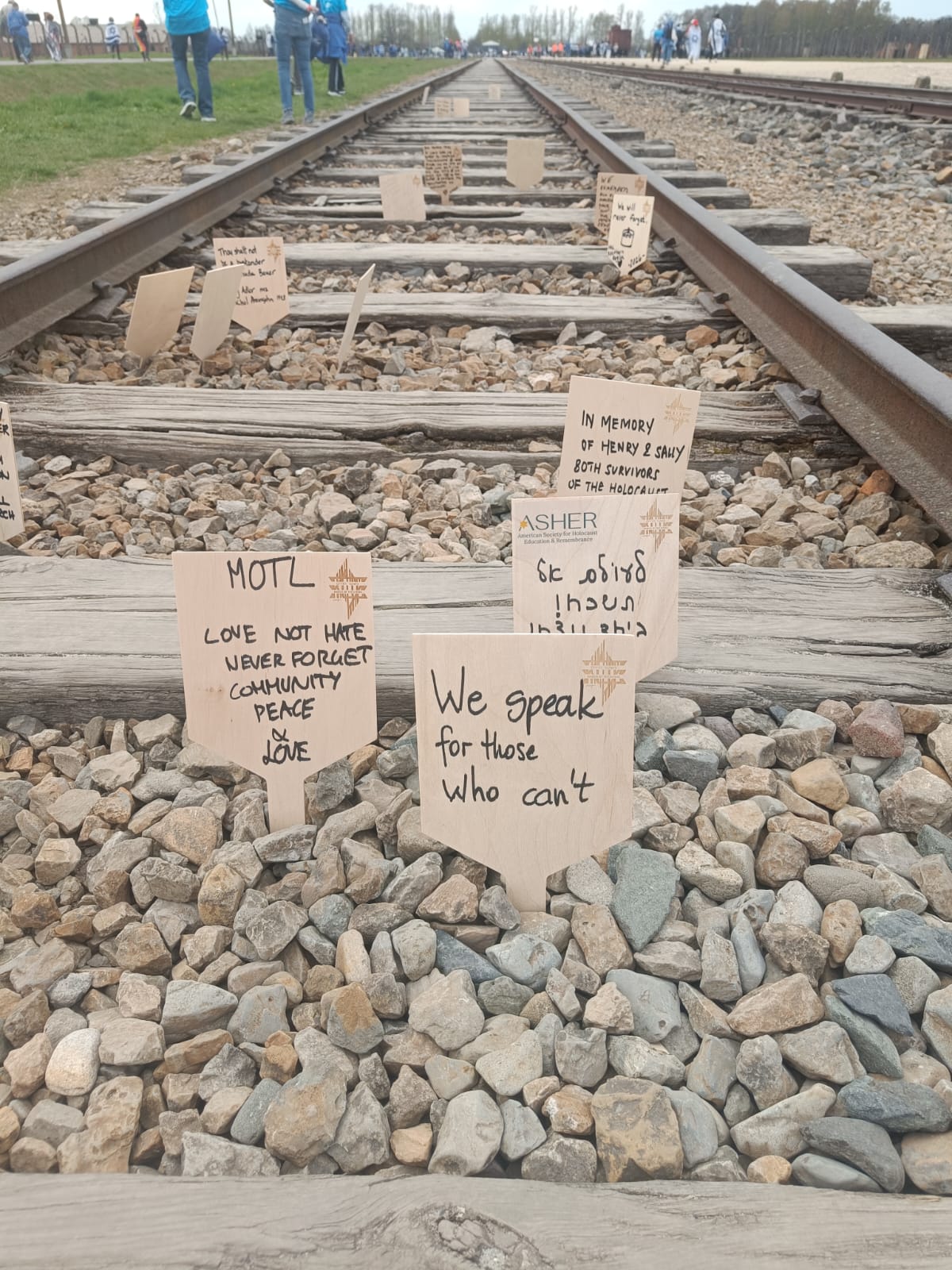 Das Bild zeigt eine Eisenbahnstrecke, die sich geradeaus in die Ferne zieht. Zwischen den Holzschwellen liegen viele kleine Steine, in die zahlreiche kleine Holztafeln gesteckt wurden. Auf diesen Tafeln stehen handgeschriebene Botschaften, die an Opfer des Holocaust erinnern oder für Werte wie Liebe, Frieden und Erinnerung stehen. Einige Schilder tragen englische Texte wie „We speak for those who can’t“ oder „Love not hate“.  Im Hintergrund sind Menschen zu sehen, die entlang der Gleise gehen oder stehen. Die Szene wirkt ruhig und zugleich nachdenklich. Insgesamt vermittelt das Bild eine Atmosphäre des Gedenkens und der Erinnerung an vergangenes Leid.