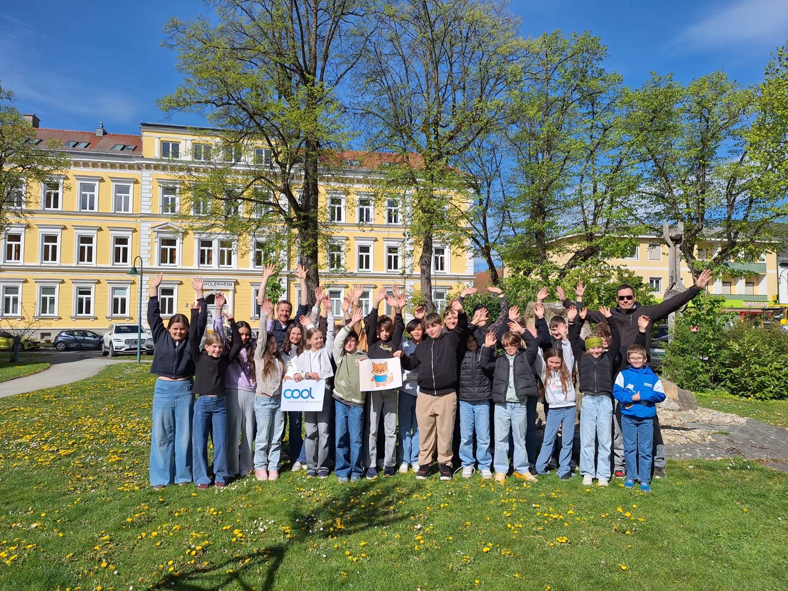 Auf dem Bild steht eine große Gruppe von Kindern und Erwachsenen auf einer grünen Wiese mit vielen gelben Blumen. Alle heben fröhlich die Arme in die Luft und schauen in die Kamera. Im Hintergrund sieht man ein großes gelbes Gebäude mit vielen Fenstern sowie hohe Bäume mit frischen grünen Blättern. Der Himmel ist strahlend blau und das sonnige Wetter sorgt für eine freundliche und lebendige Stimmung. Einige Kinder halten Schilder in den Händen, wodurch das Bild wie ein Gruppenfoto bei einer gemeinsamen Aktion oder Veranstaltung wirkt.