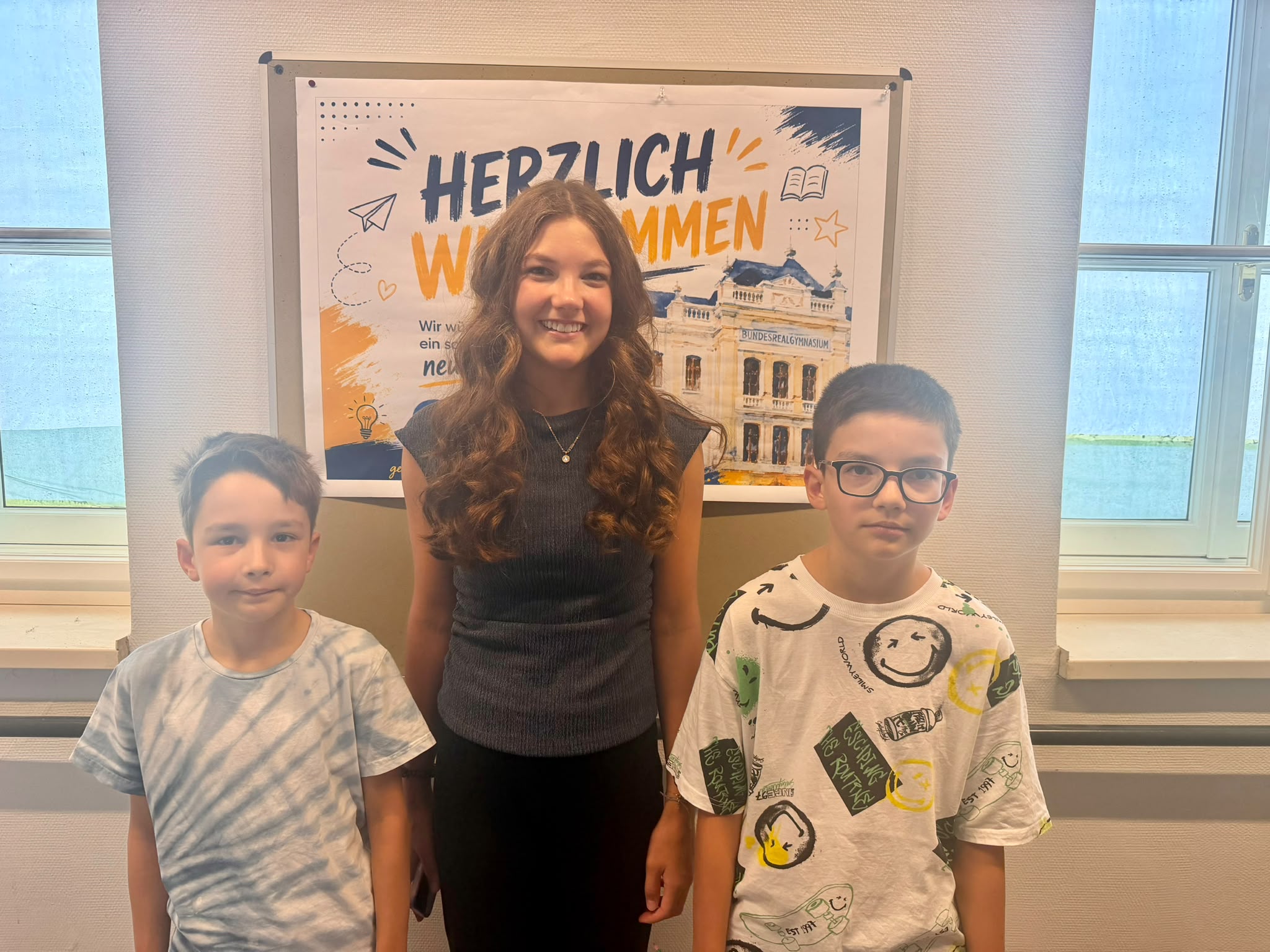 ### Bildbeschreibung  Auf dem Foto sind **drei Schülerinnen bzw. Schüler** zu sehen, die nebeneinander vor einem Plakat stehen. In der Mitte steht eine junge Frau mit langen, lockigen braunen Haaren. Sie trägt ein dunkles, ärmelloses Oberteil und lächelt freundlich in die Kamera.  Links und rechts von ihr stehen zwei jüngere Buben. Der Bub links trägt ein hellblaues T-Shirt und schaut mit einem leichten Lächeln in die Kamera. Der Bub rechts trägt eine auffällige weiße T-Shirt mit bunten Smileys und Schriftzügen und eine schwarze Brille.  Im Hintergrund hängt ein großes Plakat mit der Aufschrift **„HERZLICH WILLKOMMEN“** und einer Abbildung des **Bundesrealgymnasiums**. Das Foto wurde offenbar in einem Schulgebäude aufgenommen. Durch die Fenster an beiden Seiten fällt Tageslicht. Die drei Personen wirken freundlich und entspannt.