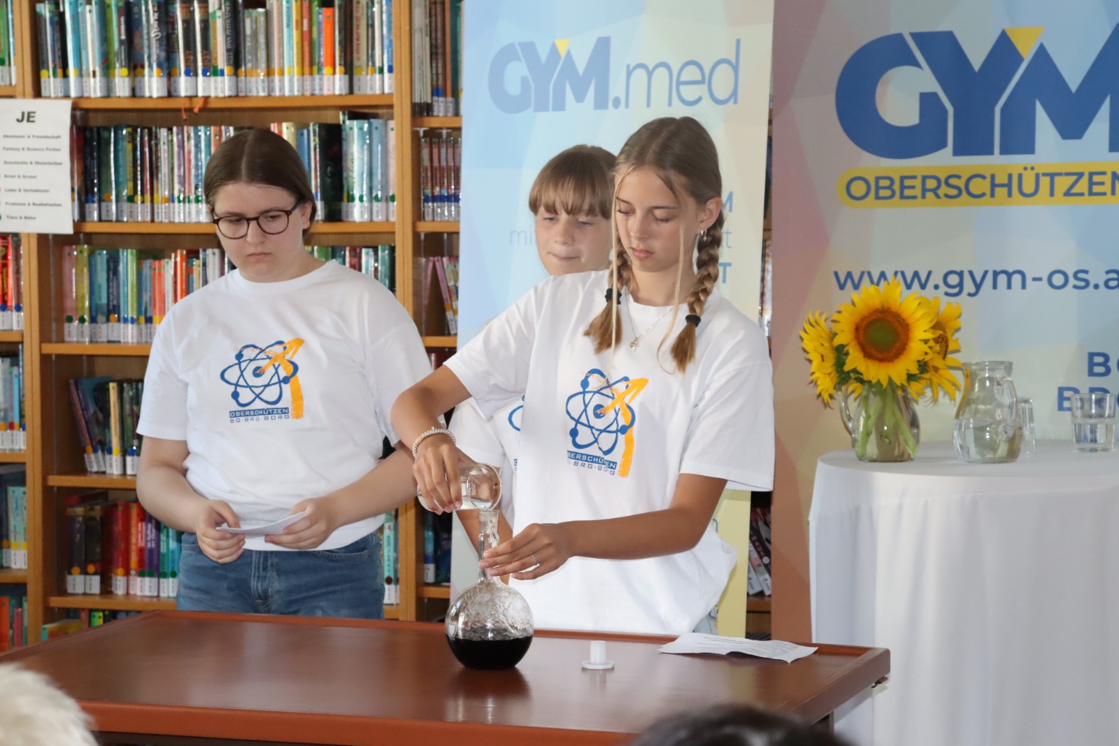 Auf dem Foto sind drei Schülerinnen bei einer schulischen Veranstaltung zu sehen. Sie tragen weiße T-Shirts mit dem Logo des Gymnasiums Oberschützen. Im Vordergrund steht ein Tisch, auf dem sich eine Glasflasche mit einer dunklen Flüssigkeit sowie weitere Utensilien befinden. Eine Schülerin gießt gerade eine Flüssigkeit in die Flasche, während die anderen beiden aufmerksam dabei sind.  Im Hintergrund sind Bücherregale einer Schulbibliothek und mehrere Roll-ups mit dem Logo des Gymnasiums Oberschützen zu sehen. Auf einem Tisch stehen außerdem ein Glasgefäß mit Sonnenblumen und mehrere Wassergläser. Insgesamt vermittelt das Bild den Eindruck einer **schulischen Präsentation oder eines naturwissenschaftlichen Experiments**.