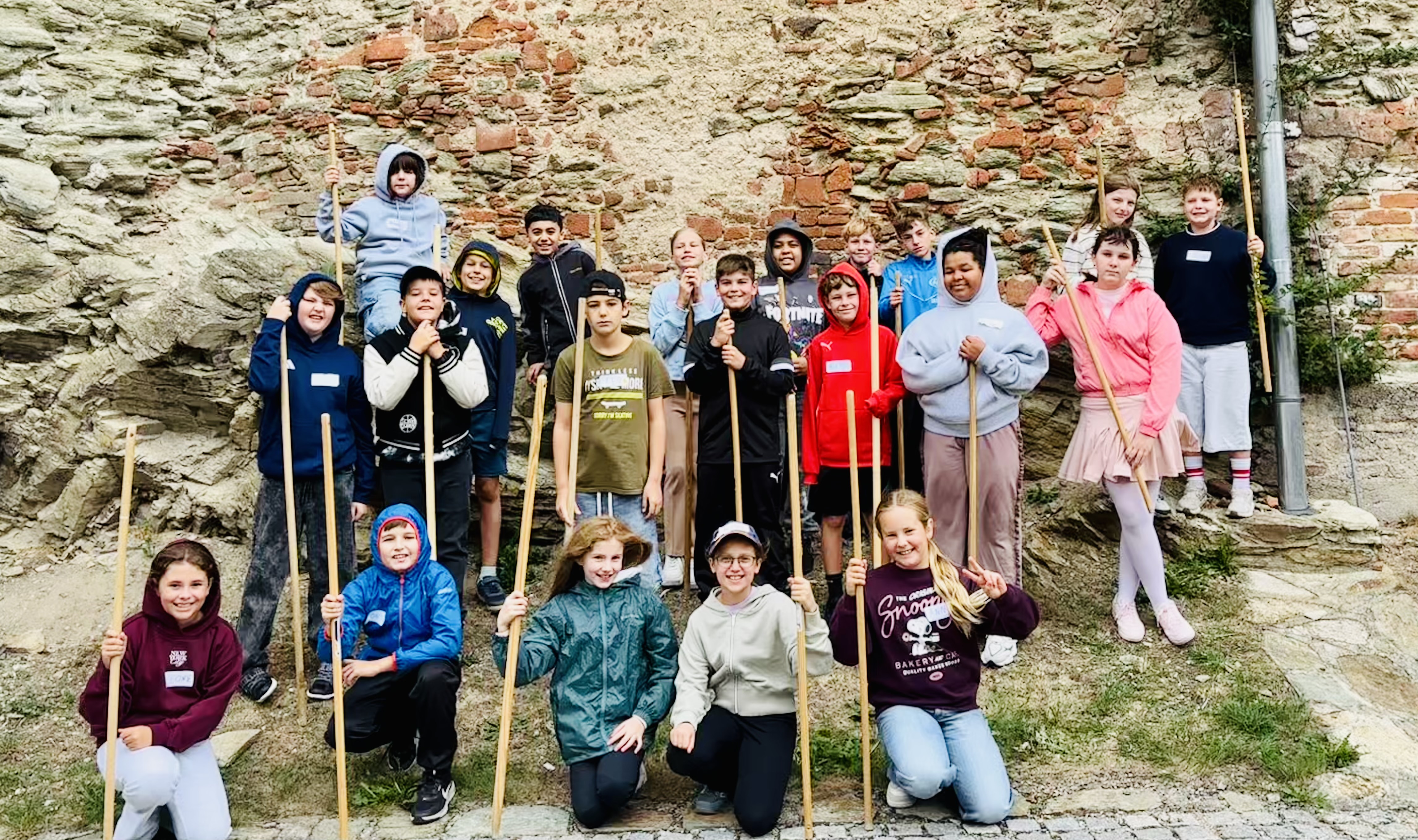 ### Bildbeschreibung  Auf dem Foto ist eine **Gruppe von Jugendlichen** vor einer alten, teilweise aus Naturstein und Ziegeln bestehenden Mauer zu sehen. Die Jugendlichen stehen und sitzen in mehreren Reihen und halten **lange Holzstäbe** in den Händen. Viele tragen wetterfeste Kleidung wie **Jacken, Kapuzenpullover und Regenjacken**, was auf einen Ausflug oder eine Outdoor-Aktivität hindeutet.  Im Vordergrund sitzen bzw. knien mehrere Jugendliche auf dem Boden. Dahinter stehen weitere Gruppenmitglieder. Die Stimmung wirkt **fröhlich und ausgelassen**. Einige Jugendliche lächeln direkt in die Kamera oder zeigen mit ihren Händen eine Geste.  Die historische Mauer im Hintergrund besteht aus unterschiedlich großen **Steinen und roten Ziegeln** und verleiht dem Bild eine rustikale Atmosphäre. Rund um die Gruppe sind Gras, Felsen und ein steiniger Untergrund zu sehen.  Insgesamt vermittelt das Foto den Eindruck eines **gemeinsamen Ausflugs oder einer Gruppenaktivität in der Natur**, bei der die Jugendlichen sichtlich Spaß miteinander haben.