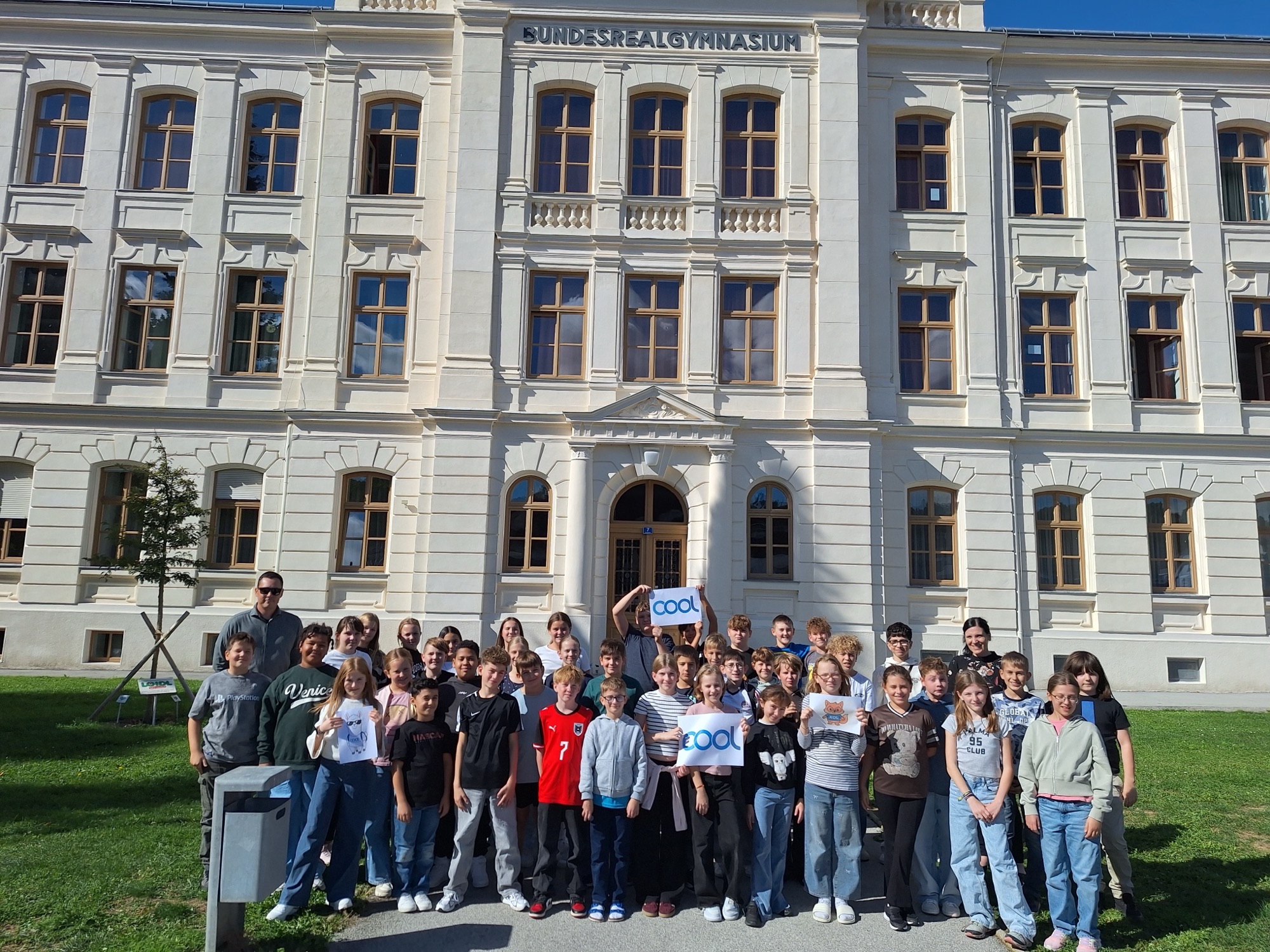 Auf dem Foto ist eine große Gruppe von Schülerinnen und Schülern gemeinsam mit zwei erwachsenen Begleitpersonen vor einem historischen Schulgebäude zu sehen. Im Hintergrund erkennt man die aufwendig gestaltete Fassade des **Bundesrealgymnasiums** mit großen Fenstern, Säulen und dekorativen Elementen.  Die Jugendlichen stehen in mehreren Reihen auf der Wiese vor der Schule und blicken in die Kamera. Einige halten Schilder mit dem **COOL-Logo** in die Höhe. Die Gruppe wirkt fröhlich und gut gelaunt. Das Foto wurde bei sonnigem Wetter aufgenommen und vermittelt eine freundliche, gemeinschaftliche Atmosphäre.
