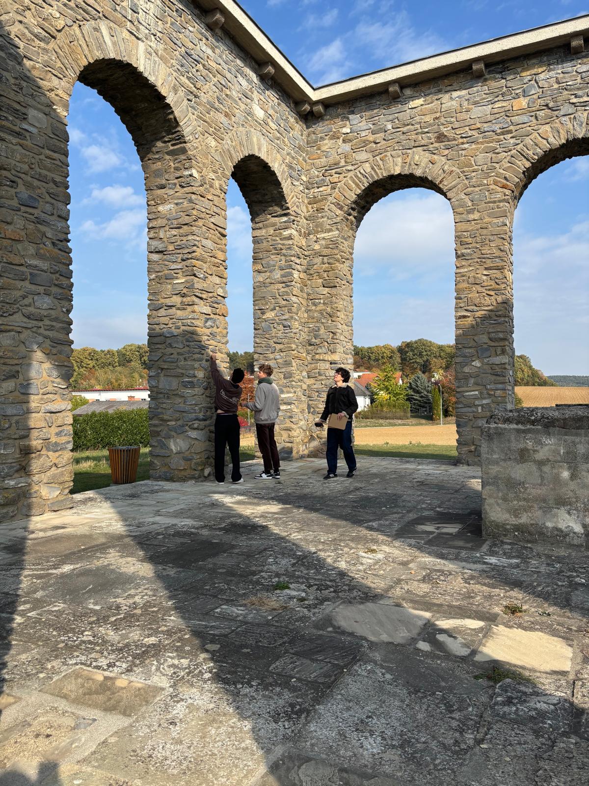 Auf dem Foto sind drei Personen inmitten einer imposanten historischen Steinbogenkonstruktion zu sehen. Die massiven Bögen und Säulen bestehen aus grob behauenen Natursteinen und verleihen dem Bauwerk einen rustikalen, historischen Charakter. Während eine Person auf einen der Steinpfeiler zeigt, betrachten die beiden anderen aufmerksam die Konstruktion. Im Hintergrund erstrecken sich grüne Wiesen, herbstlich gefärbte Bäume und eine ländliche Landschaft unter einem strahlend blauen Himmel. Das Sonnenlicht wirft markante Schatten auf den steinernen Boden und betont die beeindruckende Architektur.