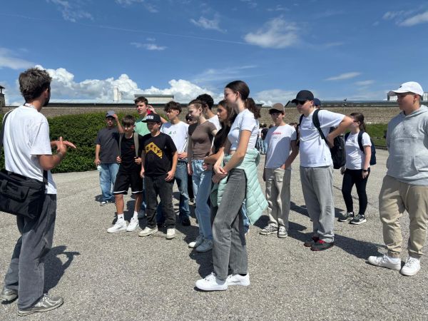Das Bild zeigt eine Gruppe von Jugendlichen und jungen Erwachsenen, die sich im Freien versammelt haben. Sie stehen auf einem gepflasterten Platz unter einem klaren, blauen Himmel mit einigen weißen Wolken. Im Hintergrund ist eine niedrige Mauer oder ein Gebäude zu sehen, das von einer Hecke flankiert wird.   Die Gruppe scheint aufmerksam einem Mann zuzuhören, der links im Bild steht und gestikuliert, als würde er etwas erklären oder eine Führung geben. Der Mann trägt ein weißes T-Shirt, eine graue Hose und eine Umhängetasche. Die Jugendlichen sind lässig gekleidet, viele tragen T-Shirts, Jeans und Turnschuhe. Einige haben Rucksäcke dabei, und eine Person trägt eine Jacke über den Armen. Die Stimmung wirkt konzentriert und interessiert, als ob die Gruppe an einer Führung oder einem Bildungsausflug teilnimmt.
