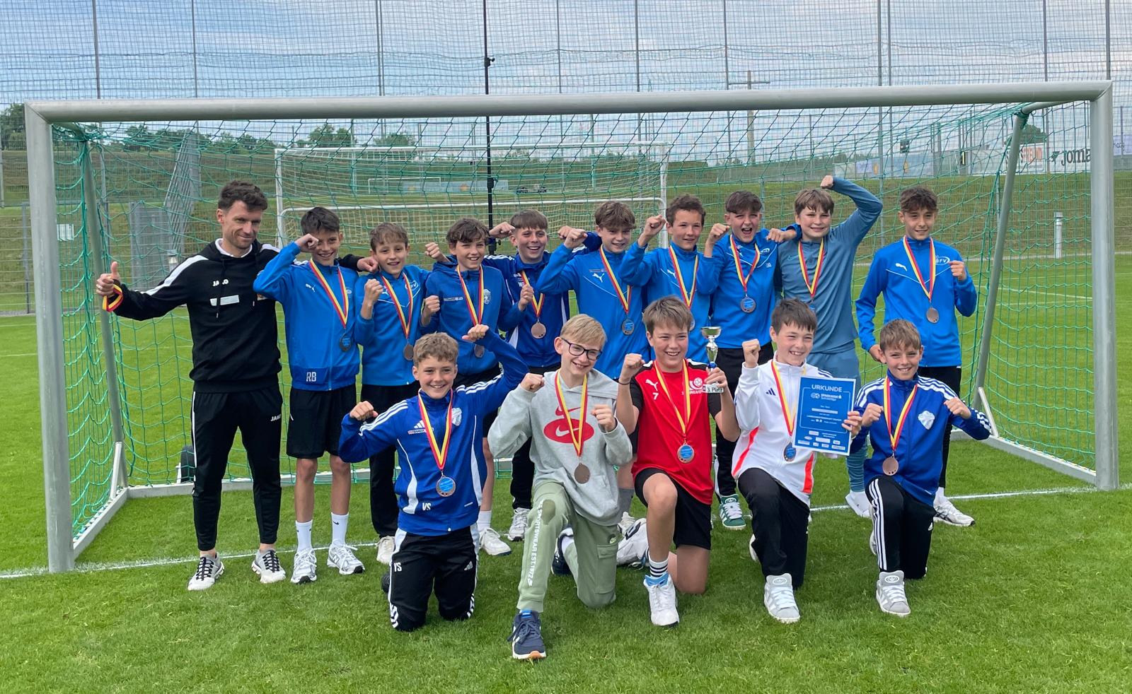 Das Bild zeigt ein Jugendfußballteam, das im Rahmen einer Schülerliga (wie der Dateiname andeutet) erfolgreich war. Die Mannschaft posiert stolz im Fußballtor auf einem Rasenplatz. Die meisten Spieler tragen blaue Trikots, während einige andere Farben wie rot und weiß tragen.  Alle Teammitglieder haben Medaillen mit farbigen Bändern um den Hals und zeigen Siegesgesten mit erhobenen Fäusten. Ein Spieler in der vorderen Reihe hält eine Urkunde oder ein Zertifikat. Links steht ein Erwachsener in schwarzer Trainingskleidung, vermutlich der Trainer des Teams.  Die Aufnahme wurde auf einem gepflegten Fußballplatz mit Kunstrasen gemacht. Im Hintergrund sind weitere Spielfelder und Fangnetze zu sehen. Die Stimmung ist fröhlich und triumphierend - ein klassisches Siegerfoto nach einem erfolgreichen Turnier.