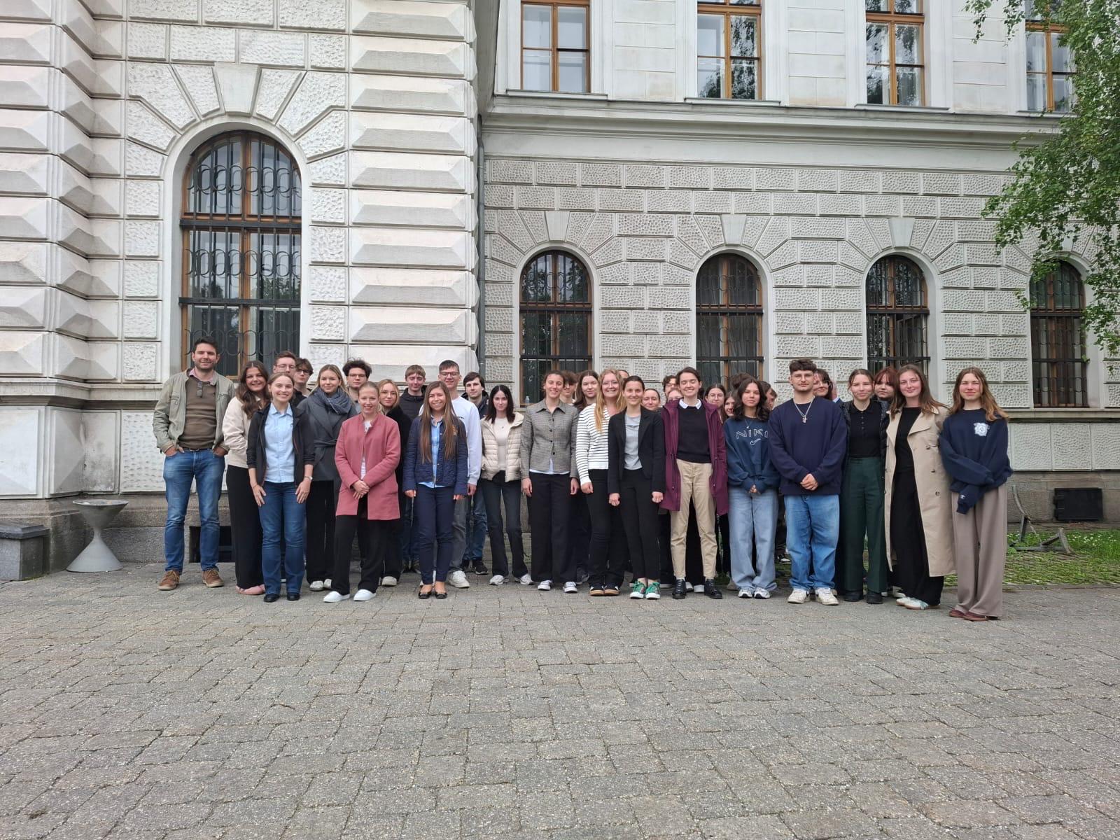 Das Bild zeigt eine große Gruppe von etwa 30-35 jungen Menschen, die für ein Gruppenfoto vor einem imposanten historischen Gebäude posieren. Das Gebäude weist eine klassische Architektur mit heller Sandsteinfassade auf, die durch charakteristische Rustikaquaderung und mehrere rundbogige Fenster mit dunklen Metallgittern geprägt ist.  Die Personen sind überwiegend Jugendliche oder junge Erwachsene, vermutlich Schüler oder Studenten, die in mehreren Reihen aufgestellt sind. Sie tragen verschiedene Freizeitkleidung - von Jeans und Pullovern bis hin zu Mäntelm und Jacken, was auf kühleres Wetter hindeutet. Die Gruppe wirkt entspannt und freundlich.  Der Schauplatz ist ein gepflasterter Innenhof oder Vorplatz mit großen Steinplatten. Im Hintergrund ist rechts ein Teil eines Baumes sichtbar, der dem Bild eine natürliche Note verleiht. Die Architektur und der Stil des Gebäudes lassen auf ein Gericht, eine Universität oder eine andere öffentliche Institution schließen, was auch zum Dateinamen &quot;Gerichtstag&quot; passen würde.