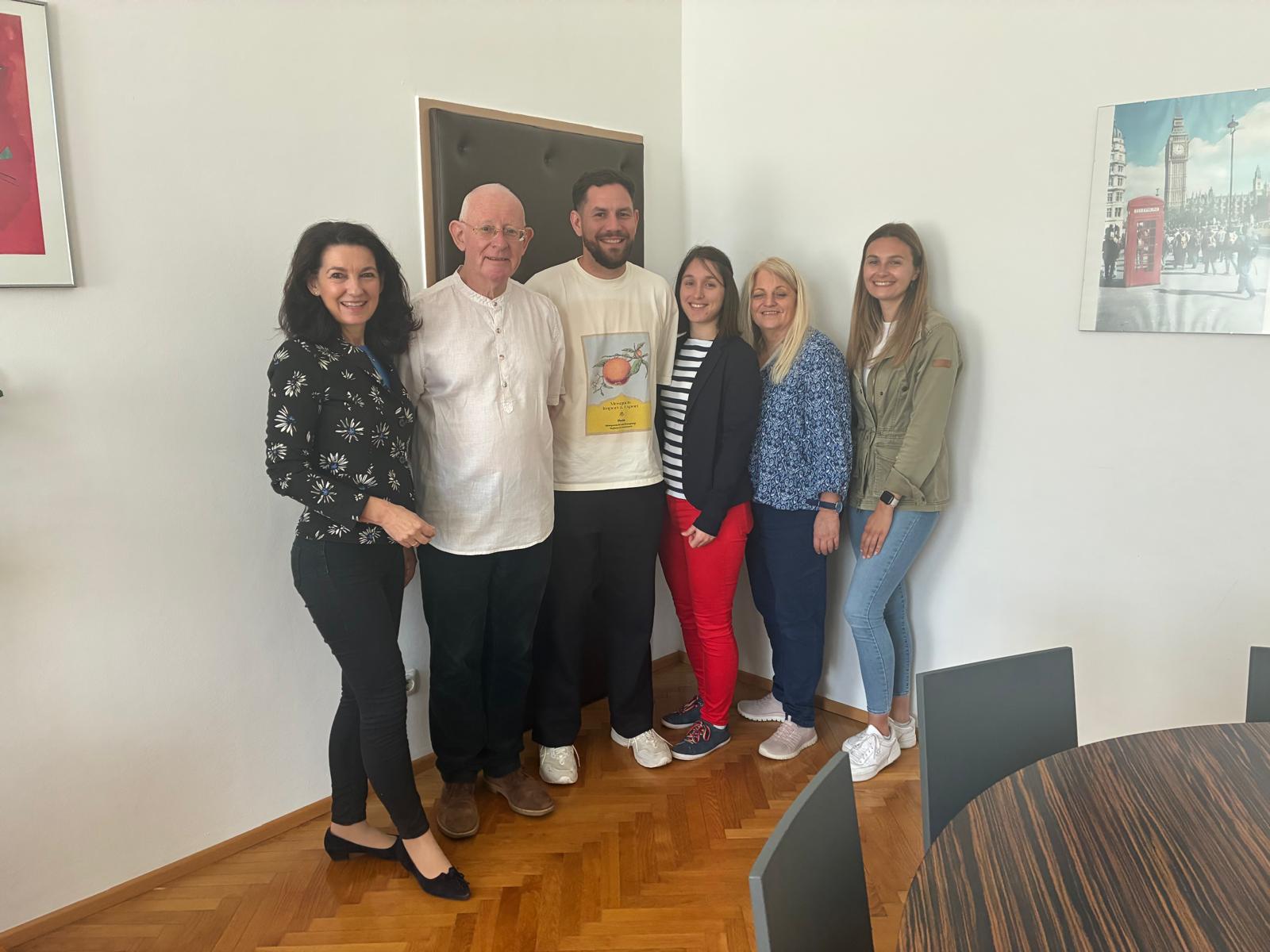 Das Bild zeigt eine herzliche Abschiedsszene in einem modernen Büro- oder Schulraum mit sechs Personen, die für ein Gruppenfoto posieren. Im Zentrum steht ein älterer Herr mit Glatze und Brille in einem weißen Hemd - vermutlich John Virgo, der Englisch-Assistent, der verabschiedet wird. Er ist umgeben von fünf Kolleginnen unterschiedlichen Alters, die alle freundlich lächeln.  Die Gruppe steht vor einer weißen Wand, an der mehrere gerahmte Bilder hängen, darunter rechts ein charakteristisches London-Motiv mit Big Ben und einer roten Telefonzelle, was perfekt zum Englischunterricht passt. Der Raum hat einen warmen Parkettboden und moderne graue Möbel sind teilweise im Vordergrund sichtbar.  Die Atmosphäre wirkt sehr herzlich und kollegial - alle Personen stehen eng beieinander und strahlen Wärme und Wertschätzung aus. Die Kleidung ist leger-professionell, von gemusterten Blusen bis hin zu Jeans und Pullovern. Das Bild vermittelt den Eindruck einer geschätzten Zusammenarbeit und eines würdevollen Abschieds von einem beliebten Kollegen.