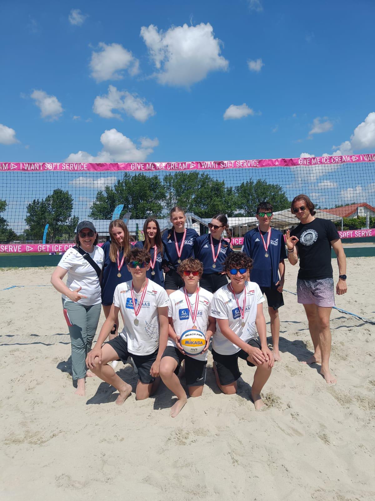 Das Bild zeigt eine erfolgreiche Beachvolleyball-Mannschaft nach einem Turnier. Elf Personen - Jugendliche und zwei Erwachsene (vermutlich Trainer/Betreuer) - posieren stolz auf einem Sandplatz vor einem Beachvolleyball-Netz.  Die Spieler tragen alle Medaillen um den Hals und sind in Teamkleidung - teils in weißen, teils in dunkelblauen Trikots. Im Vordergrund hocken drei Jungen mit Sonnenbrillen, einer hält einen Mikasa-Volleyball. Dahinter stehen die restlichen Teammitglieder und Betreuer.  Im Hintergrund ist das typische Beach-Cup-Setting zu sehen: ein strahlend blauer Himmel mit weißen Wolken, Bäume, Wohnhäuser und ein rosa Werbebanner mit &quot;GIVE ME ICE CREAM&quot; und &quot;SOFT SERVICE&quot;. Die Atmosphäre wirkt fröhlich und siegesgewiss - offensichtlich ein erfolgreicher Tag beim Beachvolleyball-Turnier.