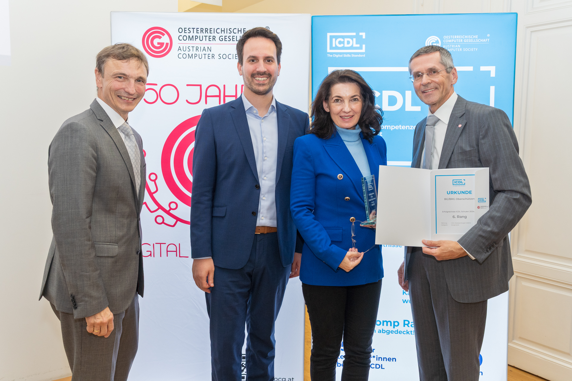 In einem hellen Raum stehen vier elegant gekleidete Personen dicht beieinander vor zwei Roll-up-Bannern. Links lächelt ein Mann im grauen Anzug in die Kamera, neben ihm ein jüngerer Mann im dunkelblauen Anzug. Rechts von ihm hält eine Frau in einem königsblauen Blazer einen gläsernen Award in den Händen. Ganz rechts präsentiert ein weiterer Mann im grauen Anzug eine geöffnete Mappe mit einer deutlich sichtbaren Urkunde, auf der „ICDL“ und „Urkunde“ zu lesen ist. Die Hintergrundbanner zeigen die Logos der Österreichischen Computer Gesellschaft und das blaue ICDL-Branding. Die Szene wirkt feierlich und vermittelt den Eindruck einer Preisverleihung im Bereich digitaler Kompetenzen; alle Beteiligten blicken zufrieden in die Kamera und präsentieren Auszeichnung und Urkunde gut sichtbar.