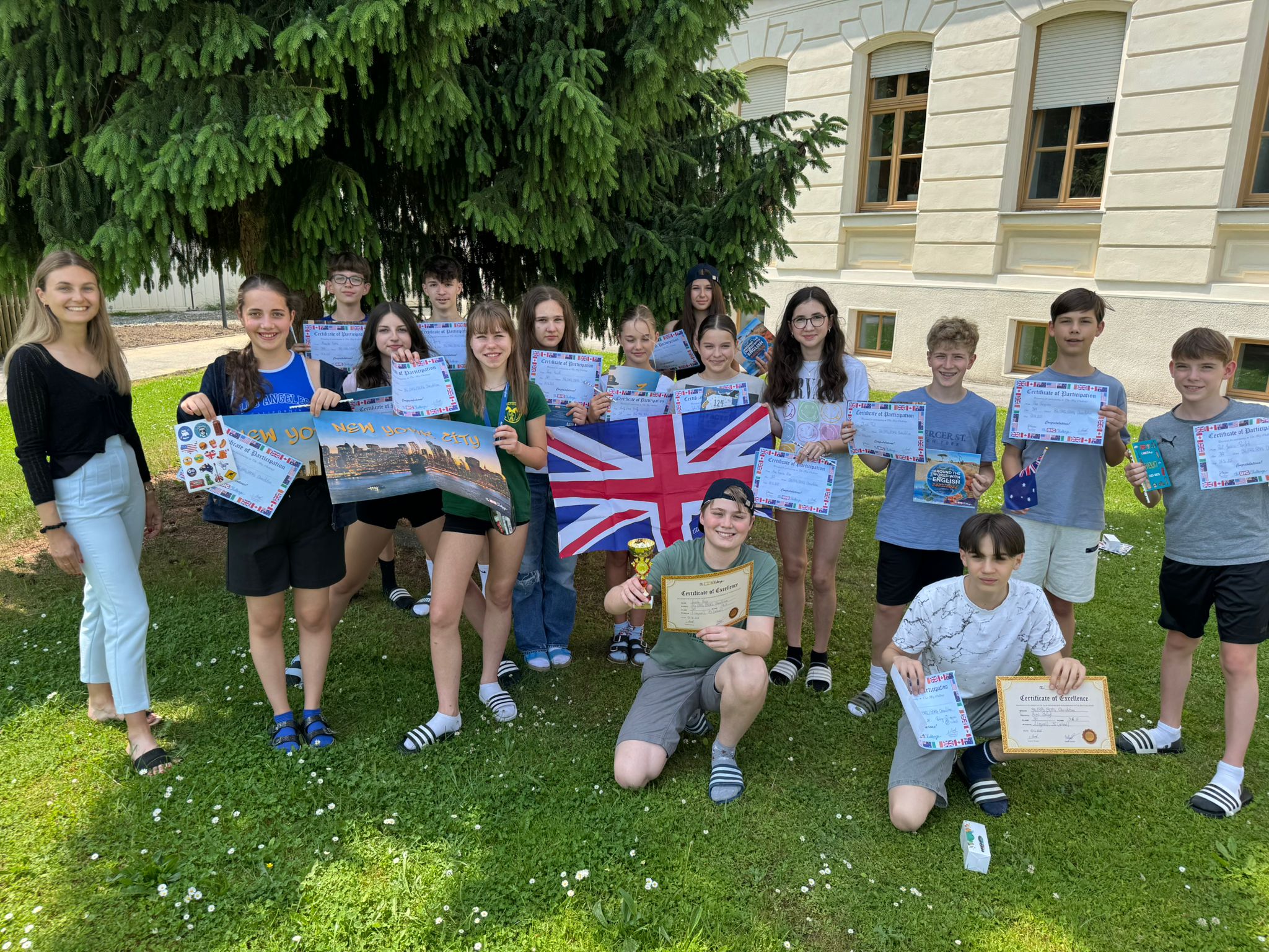 Bildbeschreibung  Eine Gruppe von Jugendlichen steht und sitzt im Freien auf einer Rasenfläche vor einem hellen, historischen Schulgebäude. Hinter ihnen ragt eine große Tanne auf. Fast alle halten bunte Zertifikate in der Hand; einige präsentieren zusätzlich Poster, kleine Preise oder eine große britische Flagge. Zwei Jugendliche knien oder sitzen vorn, einer hält einen Pokal. Links steht eine erwachsene Person lächelnd neben der Gruppe. Die Stimmung ist fröhlich und feierlich – es wirkt wie eine Preisverleihung oder der Abschluss eines Schulprojekts im Englischunterricht.