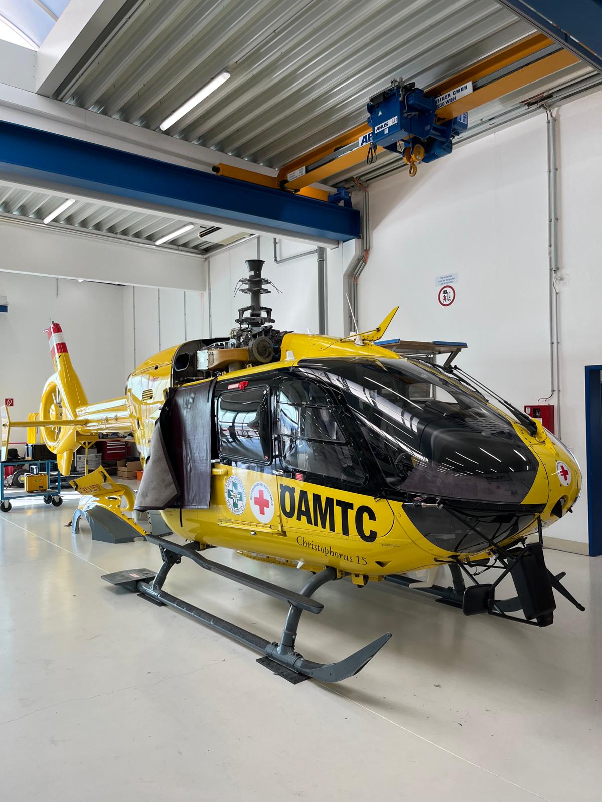 In einer hellen Wartungshalle steht ein gelber Rettungshubschrauber des ÖAMTC, seitlich mit dem Schriftzug „ÖAMTC“ und den Logos der Flugrettung versehen. Der Helikopter ruht auf grauen Kufen, die Türen sind geöffnet und geben den Blick in die Kabine frei; am Heck ist das rot-weiß gestreifte Leitwerk zu erkennen. Über dem Hubschrauber verläuft ein blauer Deckenkran, Werkstattgeräte und Werkbänke stehen im Hintergrund. Das glänzende Cockpit spiegelt das Licht der Hallenbeleuchtung, insgesamt wirkt die Szene ordentlich, technisch und betriebsbereit.