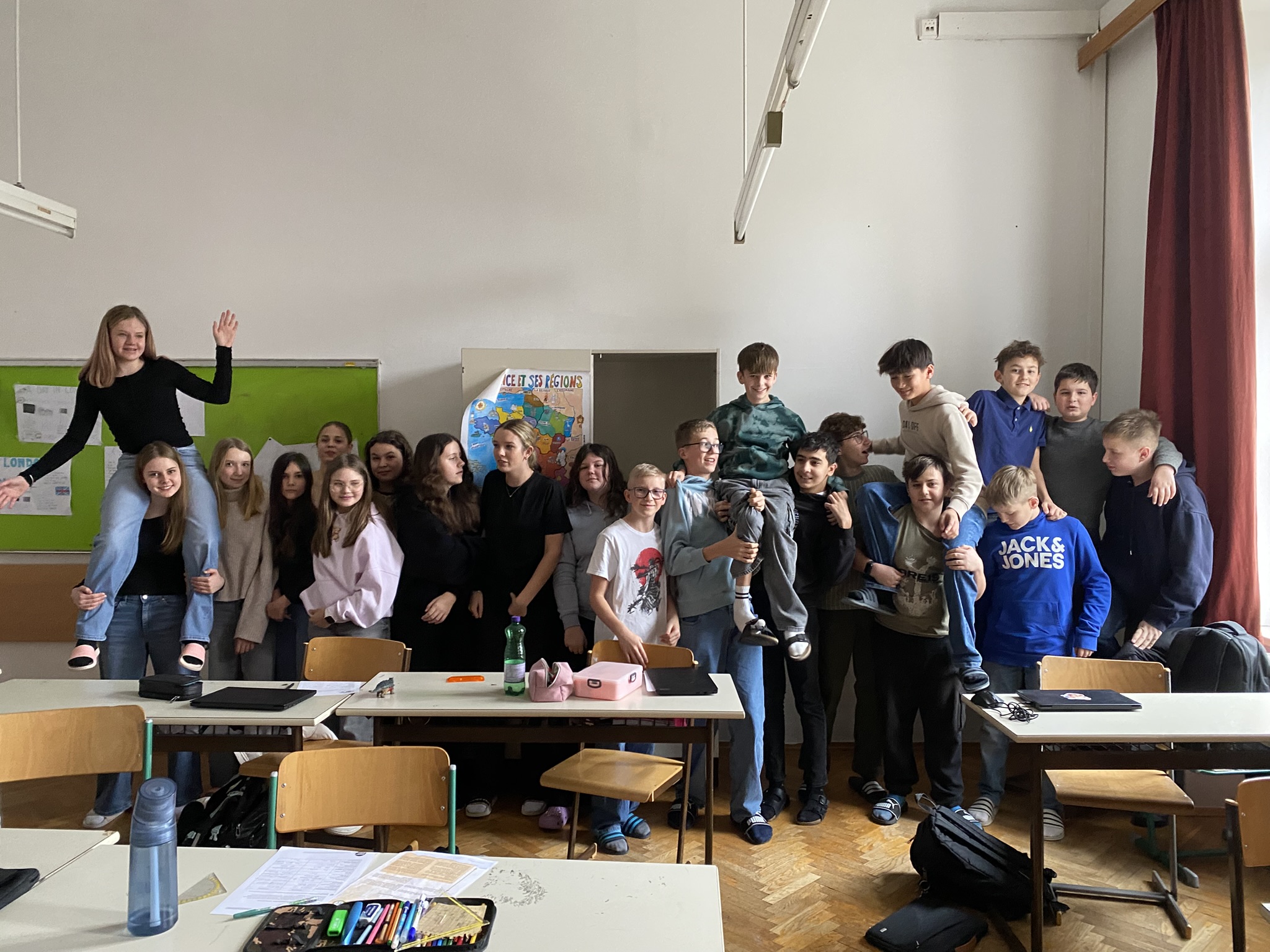 Auf dem Bild ist eine große Gruppe von Schülerinnen und Schülern in einem Klassenraum zu sehen. Die Kinder und Jugendlichen stehen dicht nebeneinander vor den Tischen und wirken fröhlich und ausgelassen. Einige von ihnen haben die Arme umeinander gelegt, andere lachen oder schauen direkt in die Kamera. Zwei Schülerinnen und Schüler sitzen jeweils auf den Schultern von Mitschülern, was der Szene eine besonders lebendige und humorvolle Stimmung verleiht.  Der Klassenraum ist hell und wirkt typisch für einen Schulalltag: Im Vordergrund stehen Tische mit Heften, Stiften und Trinkflaschen, im Hintergrund sind eine grüne Tafel sowie ein Poster an der Wand zu erkennen. Rechts hängt ein roter Vorhang vor den Fenstern. Insgesamt vermittelt das Bild ein starkes Gemeinschaftsgefühl und zeigt eine Klasse, die Spaß am gemeinsamen Lernen und Zusammensein hat.