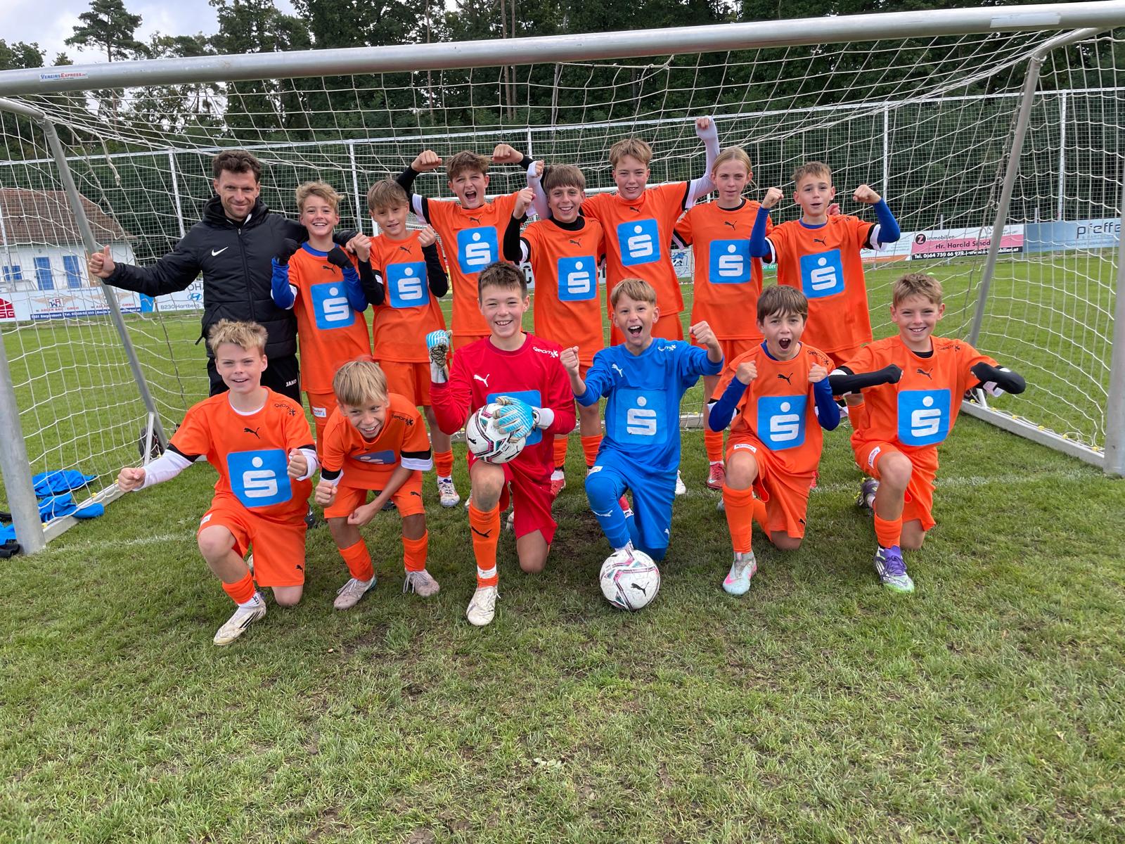 Eine fröhliche Jugendfußballmannschaft posiert vor einem Tor auf einem Rasenplatz. Die Kinder tragen leuchtend orangefarbene Trikots mit blauem Brustlogo; zwei Torhüter tragen blaue bzw. rote Torwartkleidung und halten einen Ball. Alle lächeln, jubeln und zeigen Siegerposen, einige knien im Vordergrund, andere stehen dahinter. Links steht ein Trainer im schwarzen Trainingsanzug neben dem Pfosten. Im Hintergrund sind weitere Tore, Bandenwerbung und Bäume zu sehen. Die Stimmung ist ausgelassen und sportlich.