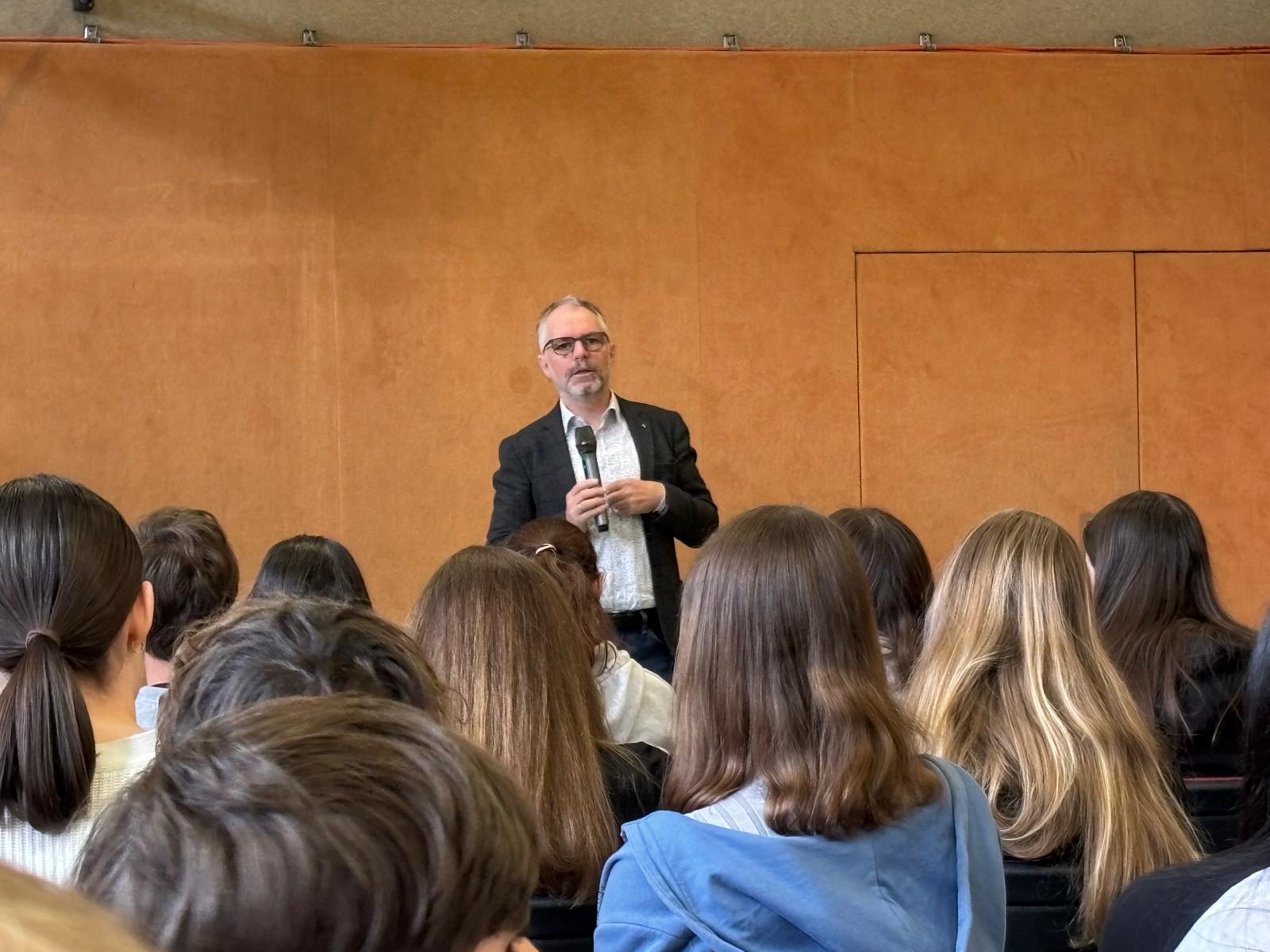 Das Bild zeigt einen Vortrag oder eine Präsentation in einem Saal mit holzvertäfelten Wänden in warmem Orangeton. Im Zentrum steht ein Redner mit Brille, der ein dunkles Sakko über einem hellen, gemusterten Hemd trägt. Er hält ein Mikrofon in der Hand und spricht zu einem Publikum.  Im Vordergrund sind die Rückenansichten zahlreicher Zuhörer zu sehen, überwiegend junge Menschen mit verschiedenen Haarfarben und -längen. Die Zusammensetzung des Publikums deutet auf eine Schulveranstaltung oder einen Vortrag für Jugendliche hin.  Der Raum wirkt wie ein Vortrags- oder Konferenzsaal mit professioneller Ausstattung. Das Bild wurde laut Dateinamen am 17. Mai 2025 aufgenommen und über WhatsApp geteilt.